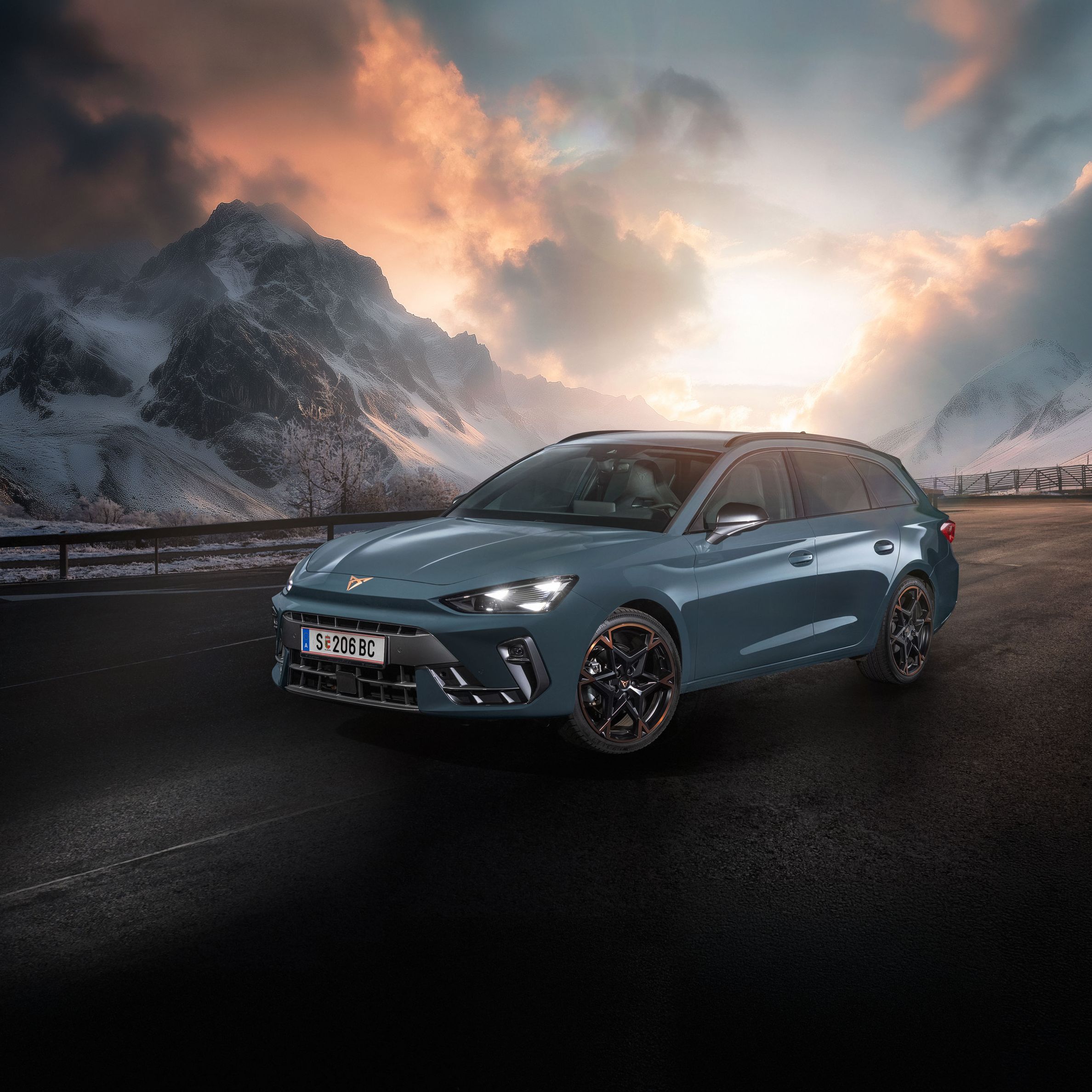Ein CUPRA Leon Kombi parkt vor einer malerischen Berglandschaft bei Sonnenuntergang. Im Hintergrund sind verschneite Gipfel und dramatische Wolken zu sehen. Der Asphalt ist dunkel und feucht.