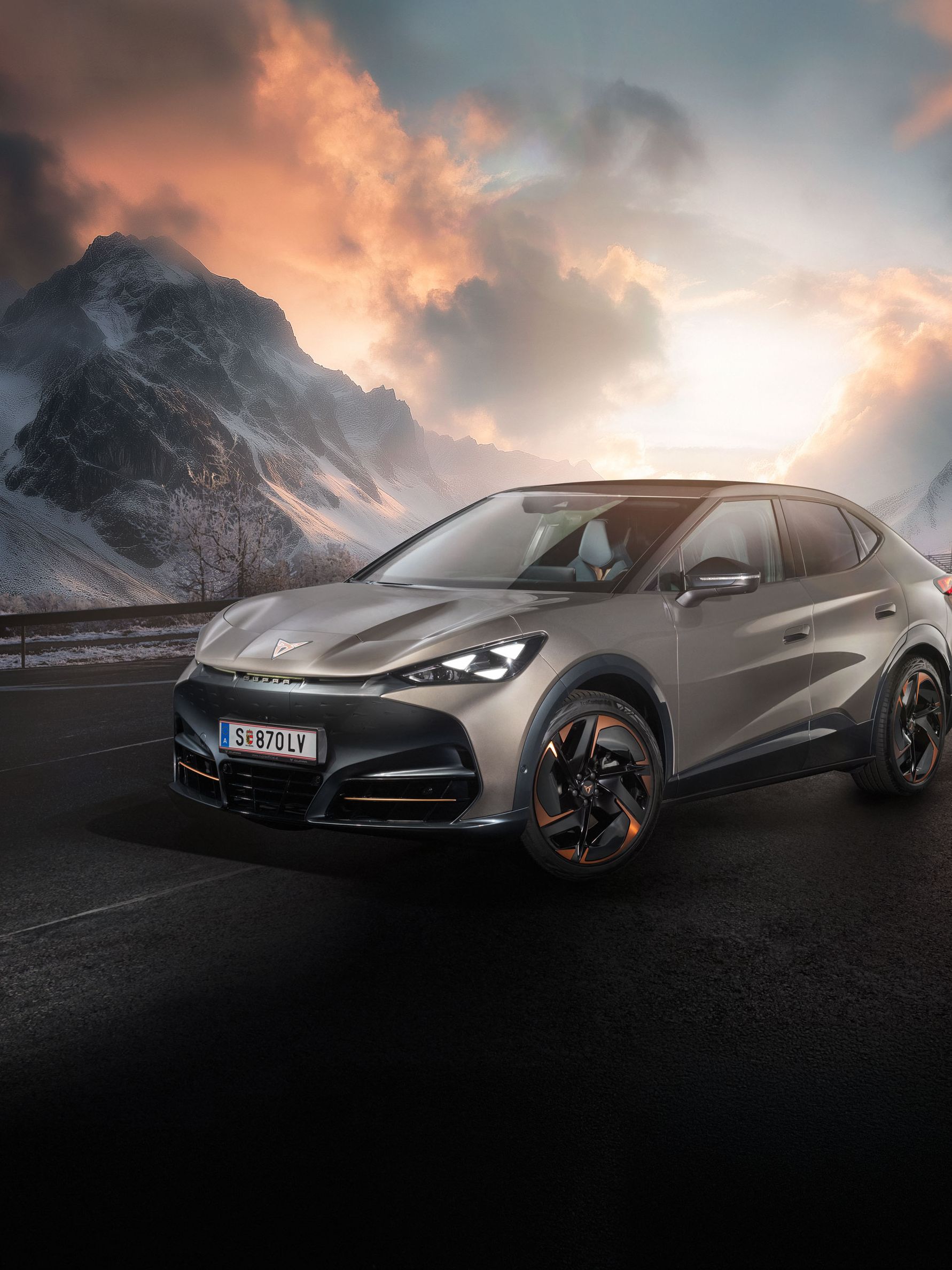 Ein CUPRA Tavascan steht auf einer Straße in einer bergigen Landschaft. Der Himmel zeigt dramatische Wolkenformationen bei Sonnenuntergang. Im Hintergrund sind schneebedeckte Gipfel zu sehen.