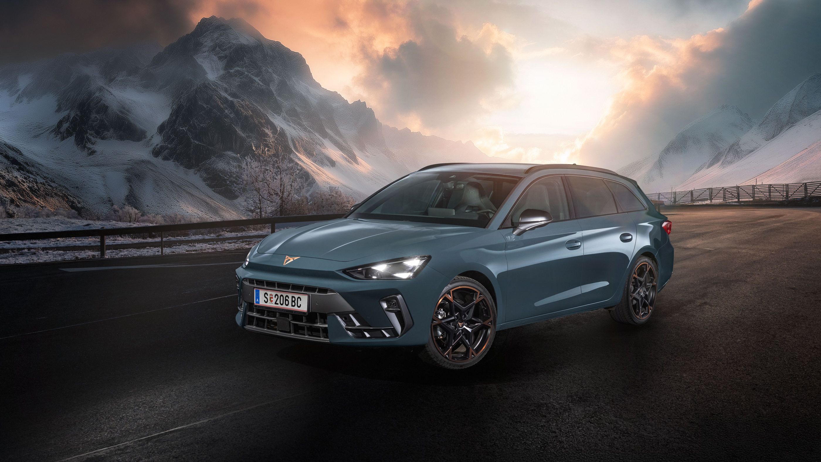Ein CUPRA Leon Kombi parkt vor einer malerischen Berglandschaft bei Sonnenuntergang. Im Hintergrund sind verschneite Gipfel und dramatische Wolken zu sehen. Der Asphalt ist dunkel und feucht.