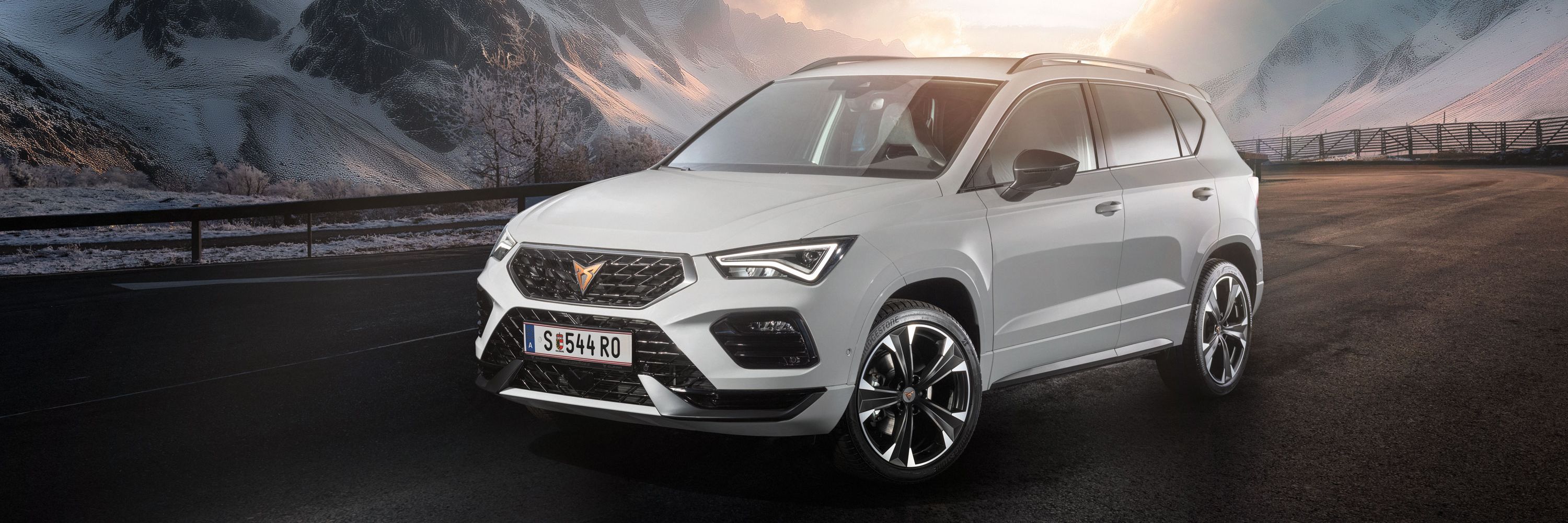 Ein weißer CUPRA Ateca fährt auf einer Bergstraße bei Sonnenaufgang. Die Straße ist nass und von Schnee gesäumt. Im Hintergrund sind schneebedeckte Berge und bewölkter Himmel zu sehen.