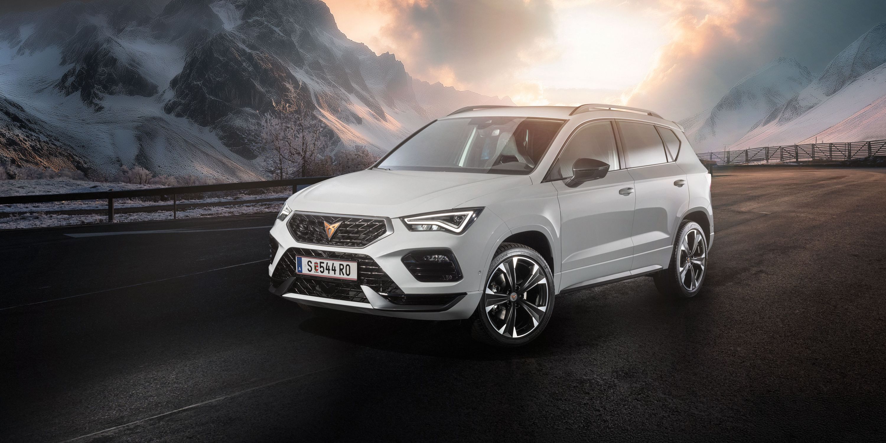Ein weißer CUPRA Ateca fährt auf einer Bergstraße bei Sonnenaufgang. Die Straße ist nass und von Schnee gesäumt. Im Hintergrund sind schneebedeckte Berge und bewölkter Himmel zu sehen.
