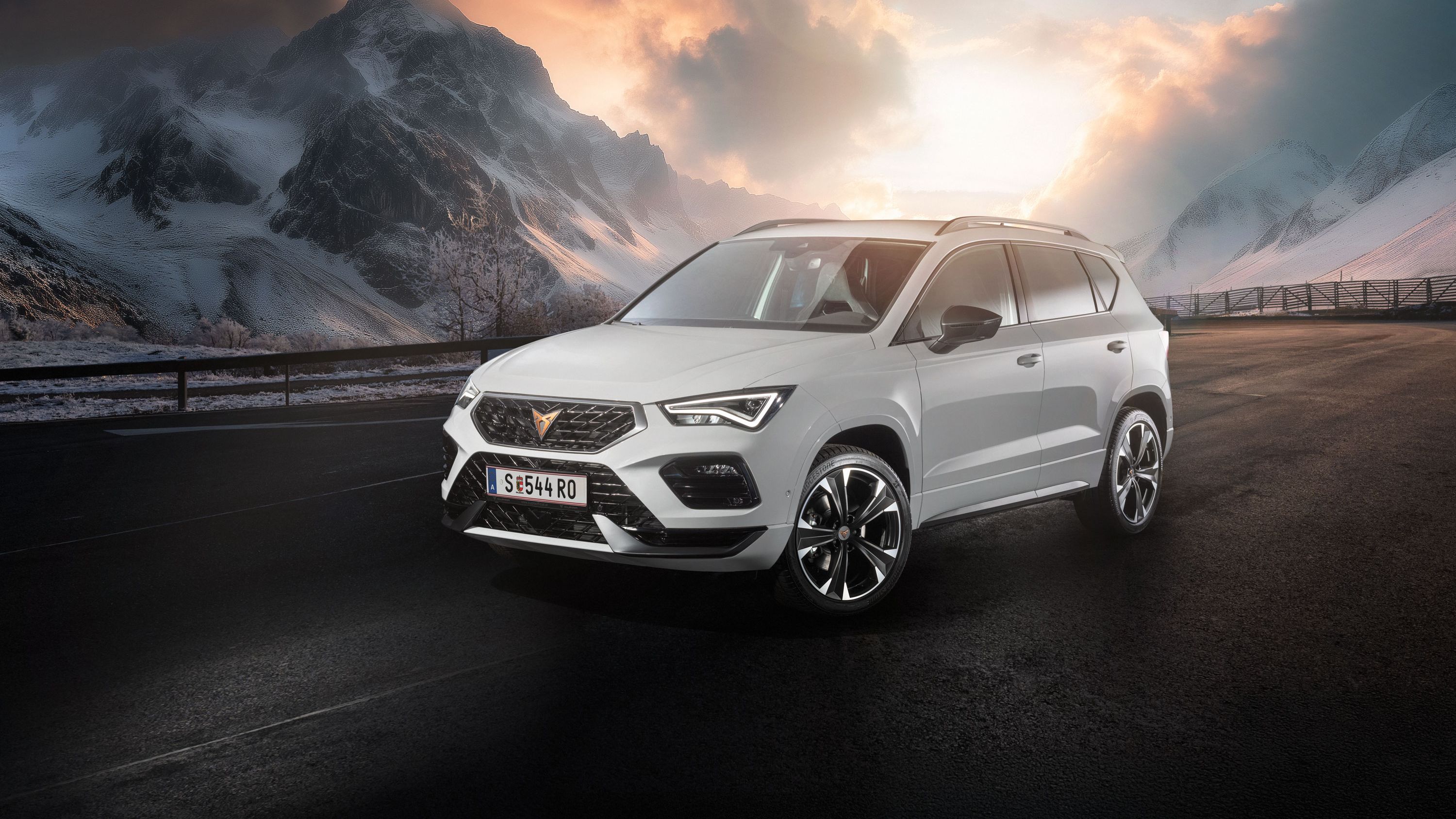 Ein weißer CUPRA Ateca fährt auf einer Bergstraße bei Sonnenaufgang. Die Straße ist nass und von Schnee gesäumt. Im Hintergrund sind schneebedeckte Berge und bewölkter Himmel zu sehen.