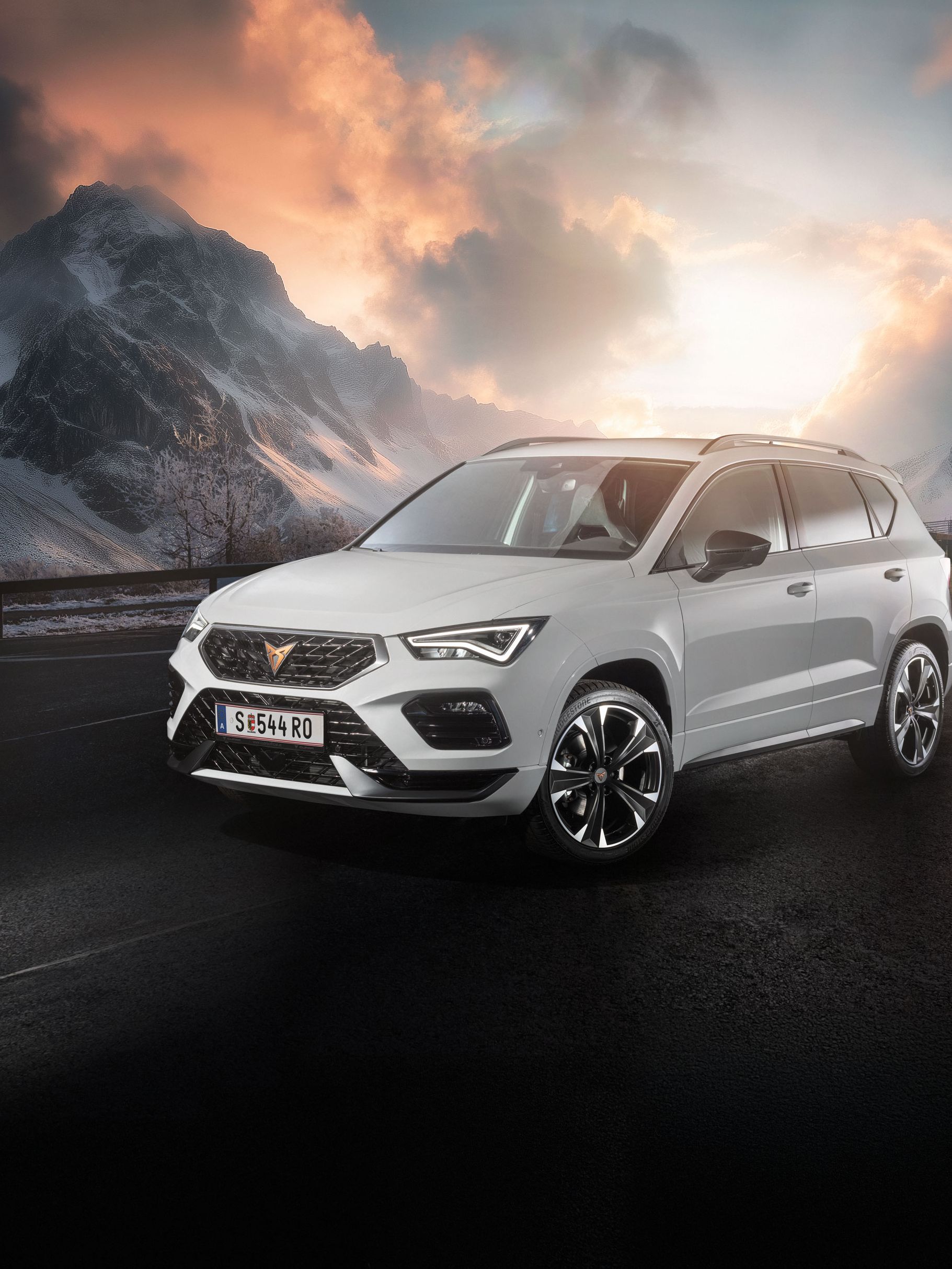 Ein weißer CUPRA Ateca fährt auf einer Bergstraße bei Sonnenaufgang. Die Straße ist nass und von Schnee gesäumt. Im Hintergrund sind schneebedeckte Berge und bewölkter Himmel zu sehen.